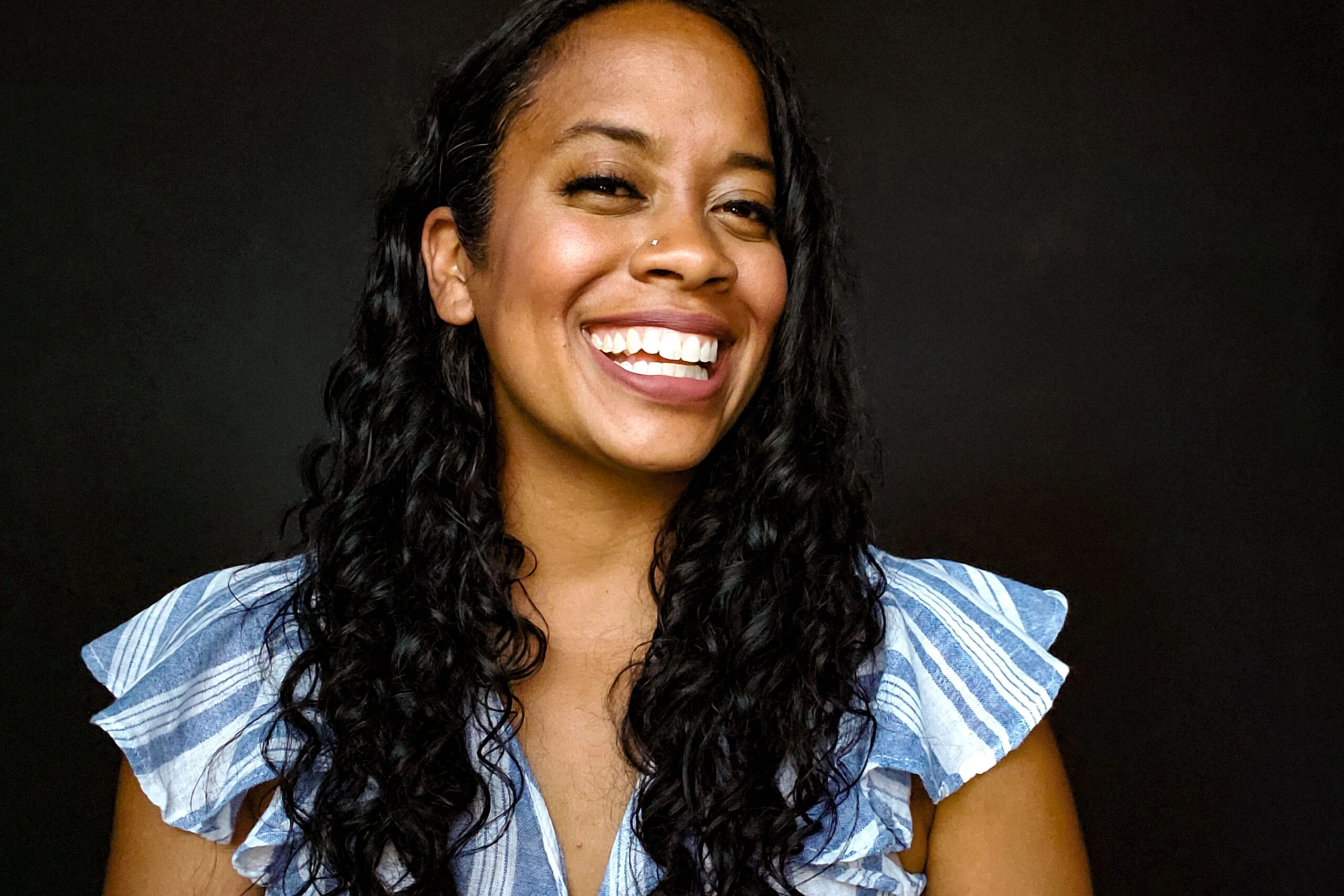 Image of Paige, a woman with long curly black hair, wearing a blue and white striped blouse, and smiling to the camera